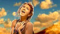 A brunette teen tilts her head upward and looks to the sky while clasping her hands at her chest and singing. She sits at the top of a red mountain cliff during sunset.