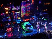 A stage with neon light signs showing a cityscape and a green car in the middle.