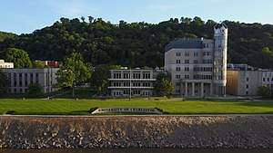 A building behind a wall that reads "University of Charleston" and another building with a glass parapet, with a river running in front