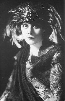 Black and white photo of a white woman wearing a feathered headdress and a dark gown with lace sleeves