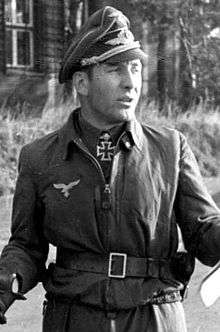 Black-and-white portrait of a man wearing a military uniform with an Iron Cross displayed at his neck, his curly hair is combed back.