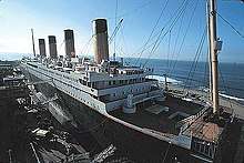 A ship resembling the Titanic is being built at a port with clear skies and small waves.