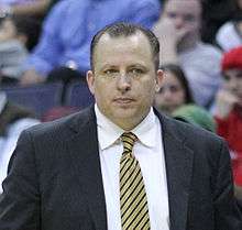 A man with dark hair, wearing a black suit, white shirt and tie, at a basketball game.