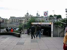 A grey, many-windowed castle with flags flying from its turrets in the background, several people walking in the foreground, and a bright sky above