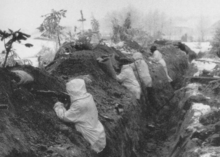 A group of soldiers with snowsuits stands in a trench, guns pointed to the left.