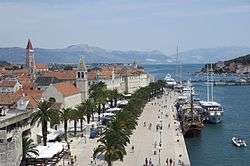 Coastal town with a palm tree promenade and houses with red roofs.