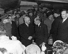 Truman and Attlee in dark suits shake hands. There are surrounded by a crowd of people, all dressed warmly. In the background is a propeller driven airliner.