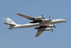 Pale grey aircraft with huge four propellers and swept back wing in flight towards right of camera.