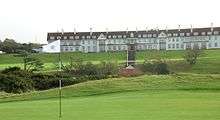  A golf course. In the background is the Turnberry Hotel, a two-story hotel with white façade and a red roof. This picture was taken in Ayrshire, Scotland.