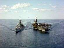 Two large warships sailing toward the viewer. On the left is a gunship, while the right ship is an aircraft carrier. A hose can be seen connecting both ships near the water line, while in the distance a helicopter can be seen in the background.