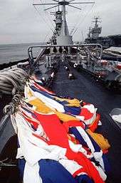 A large warship facing the viewer. In the foreground several flags of various colors can be seen on the ground. At the right of the image an aircraft carrier is visible.