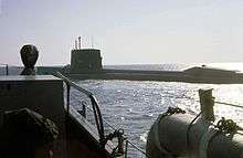 Color photograph depicting the port side of a large surfaced nuclear submarine taken from the bow of an escorting surface ship as the submarine leaves port.