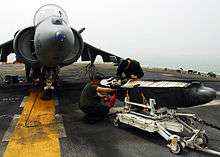 Two crew members inspecting a bomb on a trolley, in front of an aircraft.