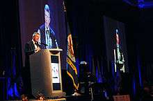 MCPON Rick D. West standing at a podium with a projection screen and two flags behind him.