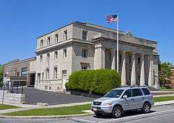 A light tan stone building with a classical colonnade and pediment on the front with an American flag flying against a blue sky from a flagpole out front. Parked on the street in front is a light blue Honda minivan.