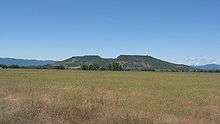 Upper Table Rock from across a field of grass, looking into the central bowl. It rises steeply from the surrounding valley to its flat top.