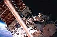 A man dressed in a spacesuit seen crawling along a white, cylindrical space station module. A large solar array can be seen projecting from the top of the module, and various other pieces of apparatus are visible. The Earth's horizon and space are visible behind the solar array.