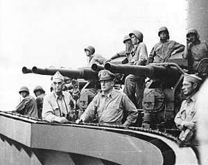 Sailors wearing steel helmets stand by an anti-aircraft gun on a quadruple mount. Two officers lean on the railing, staring off into the distance.