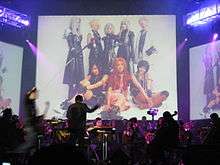 An orchestra performs a concert in a musical theatre, featuring music from the video game; in front of a screen showing people dressed as characters from the game.