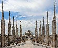 A stone rooftop, sloping down from a level center to edges where intricately decorated stone spires rise, with statues atop, proceeding toward a rear wall with some more spires. Near the walls people are gathered. Above is a blue sky largely covered with thin clouds