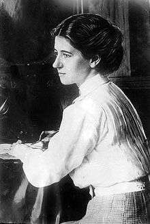 A Caucasian female sitting at a desk holding a pen, with her face in profile. She is wearing a white high collared long sleeved shirt, belt, and checkered bottom with dark upswept hair.