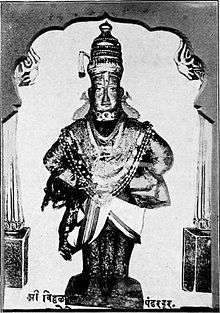 A black and white photograph of a stone icon of an arms-akimbo man standing on a brick and wearing a dhoti, angarkha (shirt), uparna (cloth that flows over both his hands), a crown, fish-shaped earrings and long necklaces till his waist.