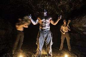 Three topless men stand in a cave.