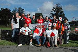 A group of thirteen supporters pose together, some wearing rugby jerseys while others sport traditional Japanese costumes and Japanese flags.