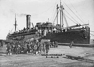 A large group of soldiers standing around a shipping dock. A ship with its gangway down is behind them.