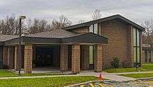 A brown rilled concrete building with a pointed roof and a similarly treated hooded entryway at left seen on a cloudy day with bare trees in the background and a small orange plastic cone on the minimal lawn in the front. On the larger block, at right, affixed capital letters spell out "Town of Wappinger"