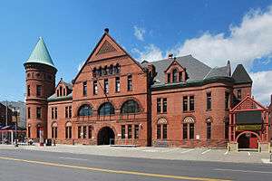 A brick building with a tall pointed roof in the center and a conical tower with light green top on the left