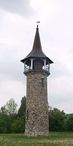 In the background is a grey, overcast sky above a canopy of trees. In the foreground is a grass field with numerous dandelions display seed heads, in the middle of which rises a tower of earth-tones multi-coloured stones. At the top of the tower is an observation deck ringed by an iron railing, each section of which is supported by end columns painted white that also support the roof structure. The copper roof is a concave structure peaking at a point, topped with an ornamental weather vane shaped like an 1800s Conestoga wagon.