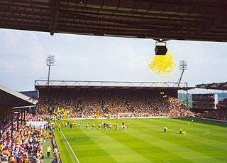 In the background and to the left are two large stands, each of which seem capable of holding thousands of people. In the foreground is a well maintained grass pitch. Yellow balloons can be seen in the sky.