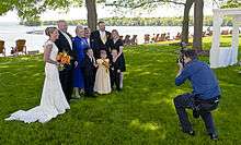 A man in a blue shirt and dark pants at right crouches as he takes a picture of a group of people at left in formal wear, with a woman in a white wedding dress at the rightmost. Behind them are tall trees and a large lake