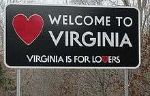 A large rectangular metal sign, mostly black, with the words "Welcome To Virginia" and "Virginia is for lovers" with a red heart symbol on the left.