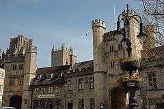 Ornate stone buildings. 2 archways beneath towers. In the foreground is a lamp stand with flowers.