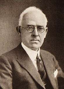 A black-and-white photo of a middle-aged Anglo man with white receding hair, round-rimmed glasses and a somewhat serious expression, wearing a dark tie and sport coat.