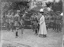 A black-and-white photograph showing a uniformed man on the left who is stooping slightly forward as he holds the hand of a man on the right, who is the king. Also in the photograph are many other uniformed men, forming a group mostly behind the two main subjects. A woman wearing a feathered hat stands to the left side of the king.