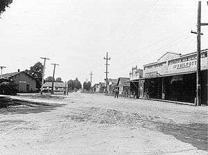 black and white photo of a street with buildings and power lines on either side and a rail line crossing lower right to mid left; station building on the left, mid frame