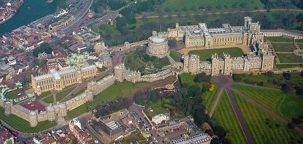 An aerial photograph of a castle, with three walled areas clearly visible, stretching left to right. Straight roads stretch away in the bottom right of the photograph, and a built-up urban area can be seen outside the castle on the left. In the upper right a grey river can just be seen.