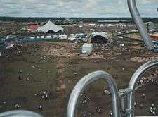 From a place up high and distant is viewed a vast open field of grass, trodden bare and brown in places with little people dotted around like ants beside enormous tents, marquees, performance stages hosting deafening arrays of musical-event speakers. The horizon is low and far away and the sky is dotted with broken clouds.