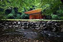 A small, boxy, wooden stage with a trapezoidal overhang stands in the center of meadow. In the foreground is a running stream with a stone embankment.