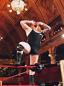 Wolfgang stands on the top turnbuckle with his hands to his mouth as he makes his signature howl taunt.