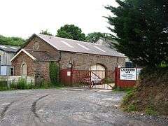 A lane, cut into the ground on the right, leads to a red fence and large single-storey stone building