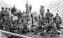 A black and white photograph of a large group of men with construction implements standing in front of the unfinished ovens