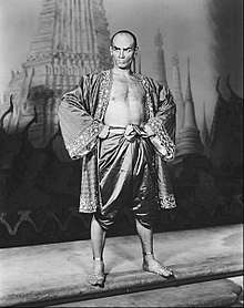 Black and white photo of a man with a shaven head in silky Asian garb; his chest is exposed and his feet are bare; he stands with hands on hips, glowering at the camera